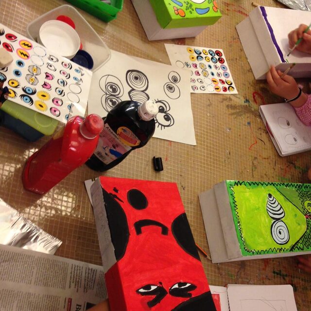 Taula de taller amb pintures, capses decorades amb cares i nens dibuixant